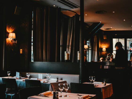 Dimly lit restaurant dining area with white-tablecloth tables, wine glasses, and candles along a dark, elegant room.