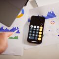 Person uses a calculator on a desk covered with financial charts, calendars, and a pencil in view.