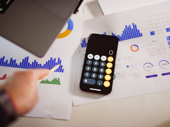Person uses a calculator on a desk covered with financial charts, calendars, and a pencil in view.