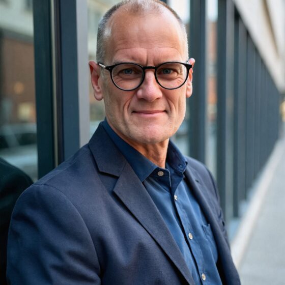 Portrait of a middle-aged man with round black glasses, wearing a navy blazer and blue shirt, beside a glass building.