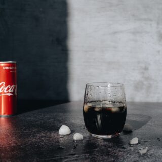 Red Coca‑Cola can on a dark countertop beside a glass of iced cola with scattered ice cubes nearby.