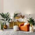 Living room corner with an orange velvet sofa, round mirror above a yellow credenza, and large green plants against a light wall.