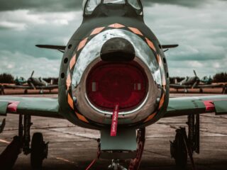 Front-on view of a vintage fighter jet with its circular intake and canopy, parked on a tarmac under a stormy sky; a red 'Remove Before Flight' tag hangs from the nose gear and a red 16 marker sits at the wheel.