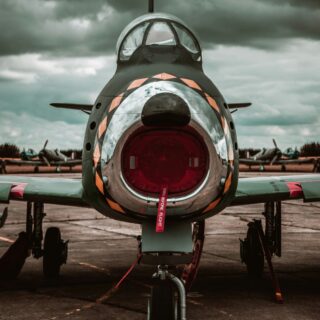 Front-on view of a vintage fighter jet with its circular intake and canopy, parked on a tarmac under a stormy sky; a red 'Remove Before Flight' tag hangs from the nose gear and a red 16 marker sits at the wheel.