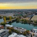 Aerial view of a city with a yellow bridge over a river, a stadium, and grid of buildings at sunset; colorful trees line the waterfront.
