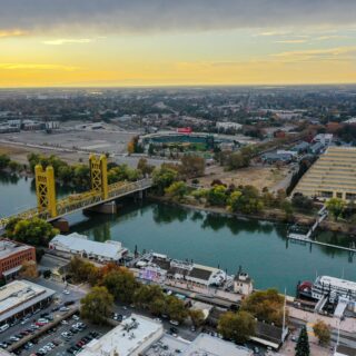 Aerial view of a city with a yellow bridge over a river, a stadium, and grid of buildings at sunset; colorful trees line the waterfront.