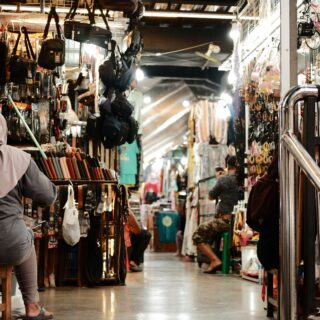 Busy indoor market stall with belts, bags, and accessories hanging along the walls as shoppers browse the aisles.