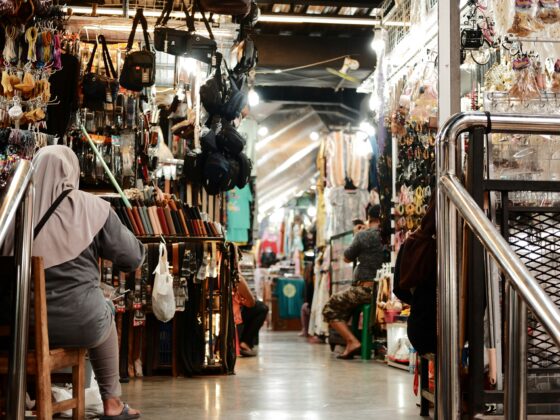 Busy indoor market stall with belts, bags, and accessories hanging along the walls as shoppers browse the aisles.