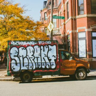 Graffiti-covered delivery truck parked on a city street, with autumn trees and red brick buildings in the background.