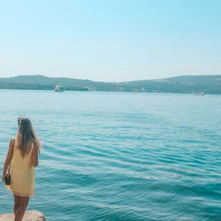 Woman in a yellow dress stands on a rocky jetty, looking out over calm turquoise water with distant hills and boats. It’s a sunny coastal scene.
