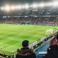 Nighttime soccer match in a packed stadium with bright floodlights and a green pitch; players on the field and banners in the stands.