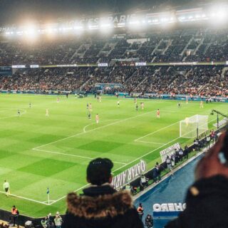 Nighttime soccer match in a packed stadium with bright floodlights and a green pitch; players on the field and banners in the stands.
