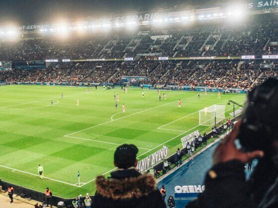 Nighttime soccer match in a packed stadium with bright floodlights and a green pitch; players on the field and banners in the stands.