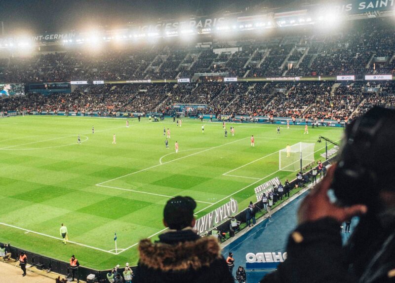 Nighttime soccer match in a packed stadium with bright floodlights and a green pitch; players on the field and banners in the stands.