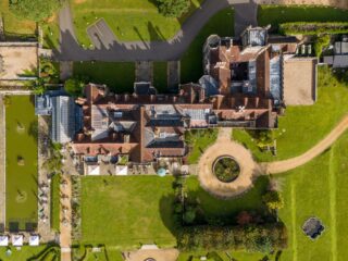 Aerial view of a historic castle with red-tiled roofs, green lawns, stone walls, and a circular gravel path with a round water feature nearby left edge.