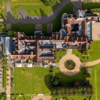 Aerial view of a historic castle with red-tiled roofs, green lawns, stone walls, and a circular gravel path with a round water feature nearby left edge.