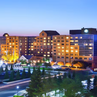 Hotel building at dusk with illuminated windows and surrounding landscaping and parking lot visible in the foreground.