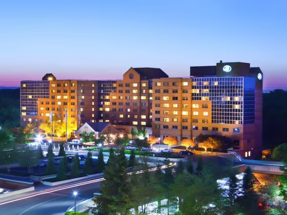 Hotel building at dusk with illuminated windows and surrounding landscaping and parking lot visible in the foreground.