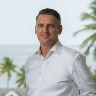 Man in a white button-down shirt smiling outdoors with palm trees and the ocean behind him in a tropical setting.