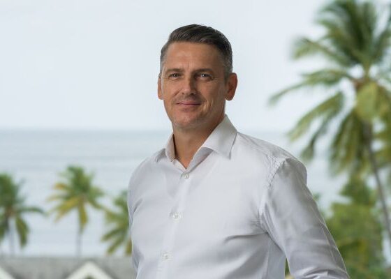 Man in a white button-down shirt smiling outdoors with palm trees and the ocean behind him in a tropical setting.