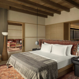 Hotel room with a king bed, white linens, a patterned headboard, pendant lights, and a seating area visible through an open doorway.