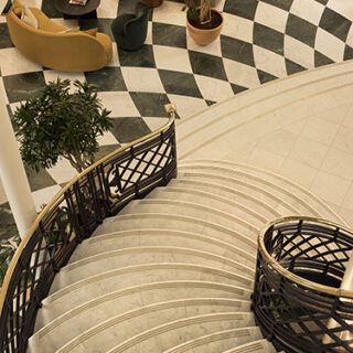 Overhead view of a curved marble staircase with a black metal railing in a lobby, visible checkered floor, seating, and potted plants nearby.