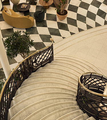 Overhead view of a curved marble staircase with a black metal railing in a lobby, visible checkered floor, seating, and potted plants nearby.