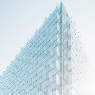 Glass office building with diagonal glass panels creating triangular, stepped patterns against a pale blue sky, viewed from the ground.