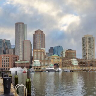 City skyline along a harbor with tall modern buildings, boats at the water's edge, and a cobblestone promenade in the foreground under a cloudy sky.