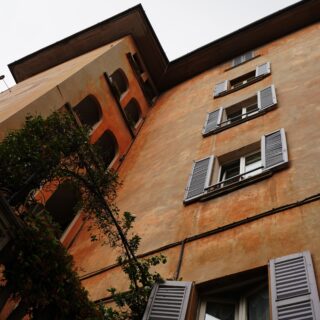 Tilted photo of an orange stucco building with white shutters and arched windows, greenery in the foreground.