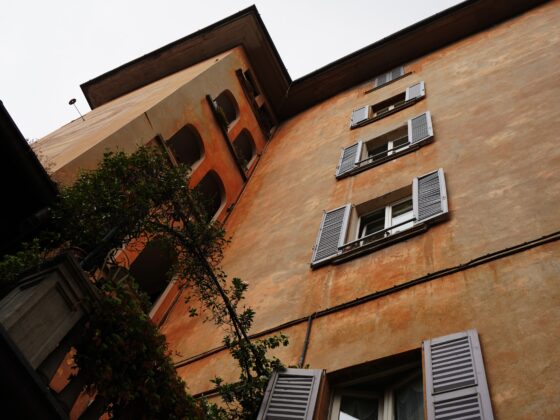 Tilted photo of an orange stucco building with white shutters and arched windows, greenery in the foreground.