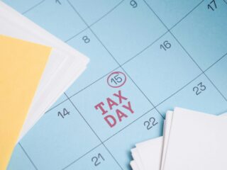Blue calendar page with red 'TAX DAY' text and a circled date 15, placed beside yellow sticky notes and white papers (informational image).