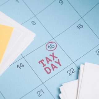 Blue calendar page with red 'TAX DAY' text and a circled date 15, placed beside yellow sticky notes and white papers (informational image).