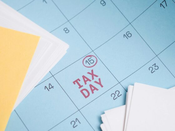Blue calendar page with red 'TAX DAY' text and a circled date 15, placed beside yellow sticky notes and white papers (informational image).