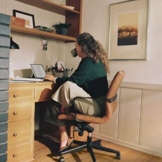 Person with curly hair seated at a wooden desk, working on a laptop with papers and supplies nearby in a cozy home office.