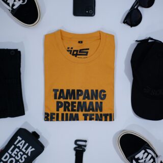 Overhead view of a clothing flat lay: a yellow T-shirt with bold black text in the center, surrounded by black jeans, sneakers, sunglasses, a belt, a black bag, and a smartphone on a white surface.