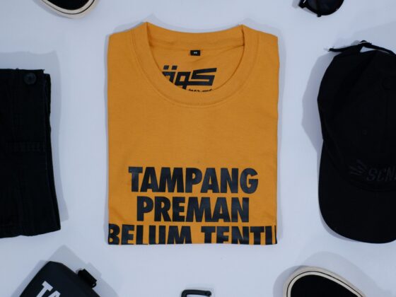 Overhead view of a clothing flat lay: a yellow T-shirt with bold black text in the center, surrounded by black jeans, sneakers, sunglasses, a belt, a black bag, and a smartphone on a white surface.