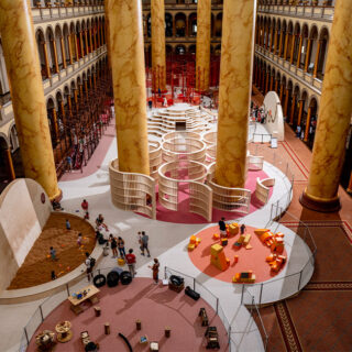 Inside a grand atrium with tall marble columns and arched balconies, a circular play area with orange seating blocks and white curved barriers dominates the center while visitors explore around it.
