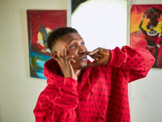 Person in a red hoodie making a playful face, pressing his cheeks with his fingers indoors, with colorful paintings in the background