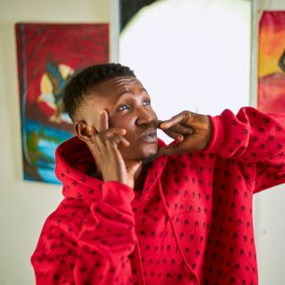 Person in a red hoodie making a playful face, pressing his cheeks with his fingers indoors, with colorful paintings in the background