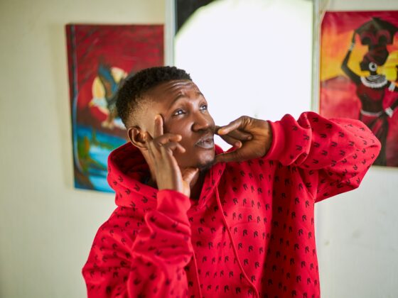 Person in a red hoodie making a playful face, pressing his cheeks with his fingers indoors, with colorful paintings in the background