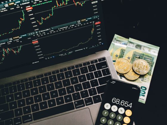 Laptop screen shows multiple financial charts and candlestick trading graphs with euro banknotes and Bitcoin coins nearby on the desk.