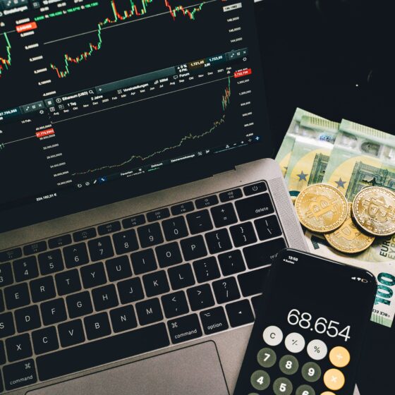 Laptop screen shows multiple financial charts and candlestick trading graphs with euro banknotes and Bitcoin coins nearby on the desk.
