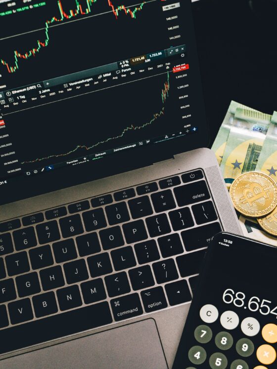 Laptop screen shows multiple financial charts and candlestick trading graphs with euro banknotes and Bitcoin coins nearby on the desk.