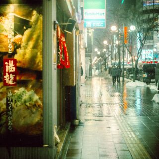 Nighttime street in Japan with a large food poster on the left, red signs, and people walking in the rain.