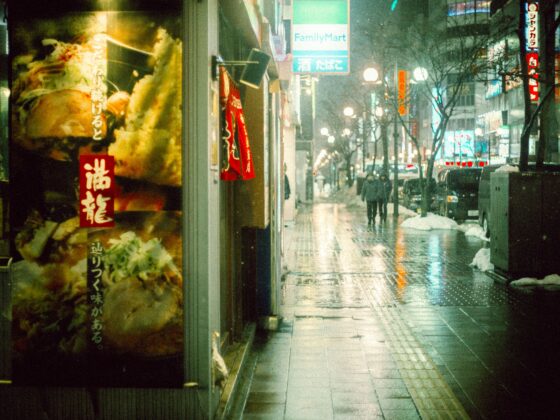Nighttime street in Japan with a large food poster on the left, red signs, and people walking in the rain.