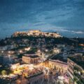 Nighttime view of the illuminated Acropolis atop a hill, with a busy market plaza and city below.