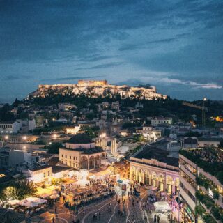 Nighttime view of the illuminated Acropolis atop a hill, with a busy market plaza and city below.