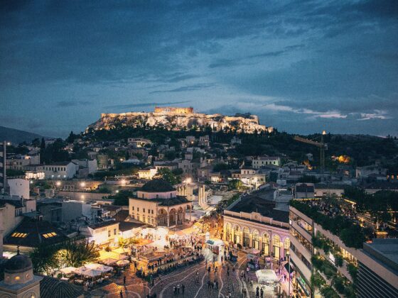 Nighttime view of the illuminated Acropolis atop a hill, with a busy market plaza and city below.