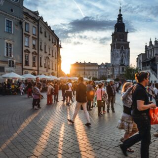 Crowded European city square at sunset with cafes and historic buildings, a tall clock tower in the background under a blue sky.